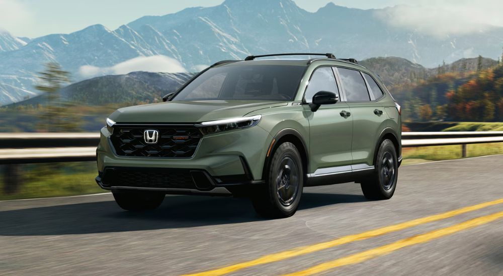 A green 2027 CR-V TrailSport Hybrid driving past a guard rail.