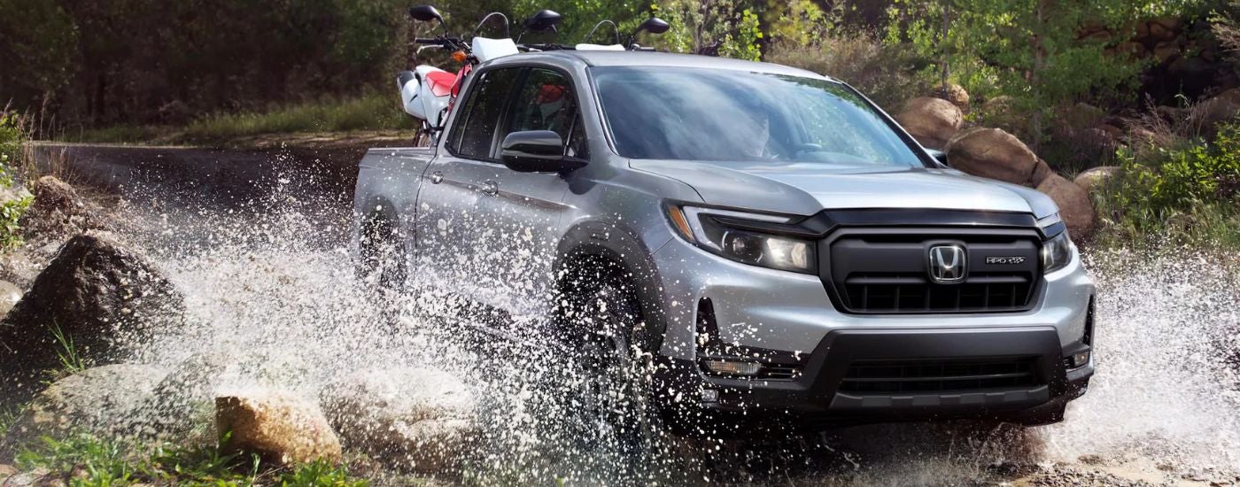 Silver 2026 Honda Ridgeline HPD with a dirt bike in the back driving through a river.